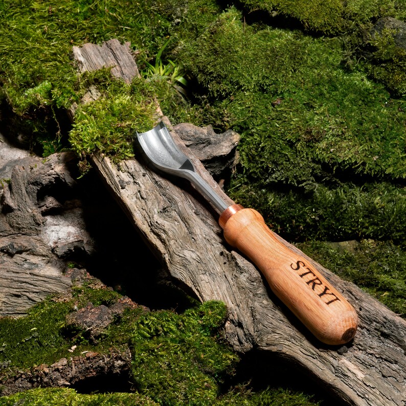 Pu&ograve; includere: Uno strumento per intagliare il legno con una lama in metallo e un manico in legno. Il manico &egrave; inciso con la parola "STRYI". Lo strumento &egrave; appoggiato su un pezzo di legno ricoperto di muschio.