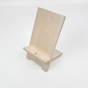 Peut inclure: Un support de téléphone en bois de couleur claire. Le support est en bois non fini et comporte un support arrière et une petite étagère avec un trou circulaire. Le support est conçu pour maintenir un téléphone ou une tablette en position verticale.