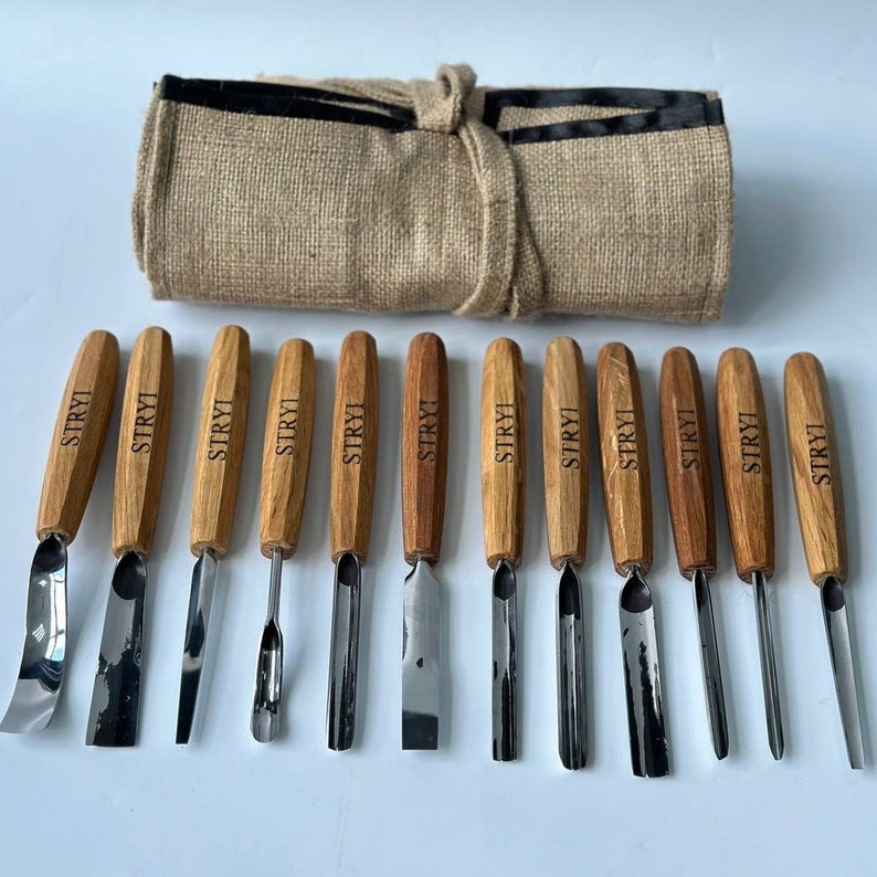 May include: A set of 12 wood carving tools with a brown leather roll-up case. The tools are made of wood and metal and have a variety of shapes and sizes. The tools are labelled "STRYI" on the wooden handles.