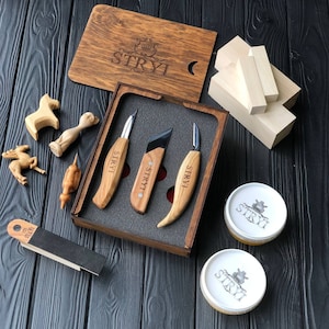 Op de afbeelding: Een set van drie houtsnijmesjes in een houten doos met een zwart schuim interieur. De messen hebben houten handvatten met het woord "STRYI" erop gegraveerd. De doos is ook gegraveerd met het woord "STRYI".