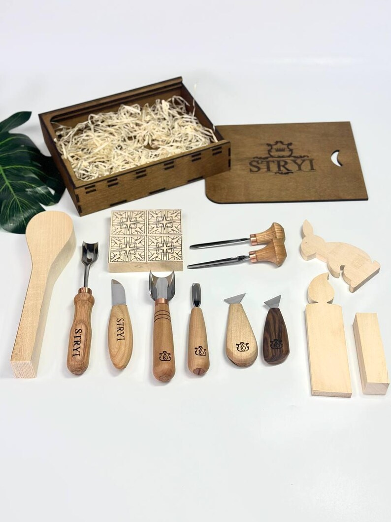 May include: A wood carving set featuring a selection of tools such as gouges, knives, and a spoon. The set includes a wooden box, and the brand name "STRYI" is visible on some tools. Wooden cutouts of a rabbit and other shapes are also included.