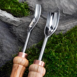 May include: Two metal carving tools with wooden handles. The tools have a curved blade and a rounded tip. The tools are on a background of green moss and gray rock.