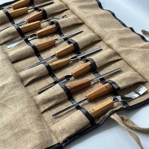 May include: A brown burlap roll-up case with black trim, containing a set of wood-handled carving tools. The tools have metal blades and are labelled "ST".