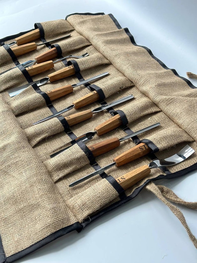May include: A brown burlap roll-up case with black trim, containing a set of wood-handled carving tools. The tools have metal blades and are labelled "ST".