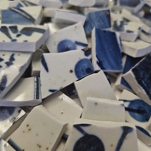 Op de afbeelding: Een stapel gebroken witte keramische tegels met blauwe en zwarte geverfde ontwerpen. De tegels zijn onregelmatig van vorm en grootte.
