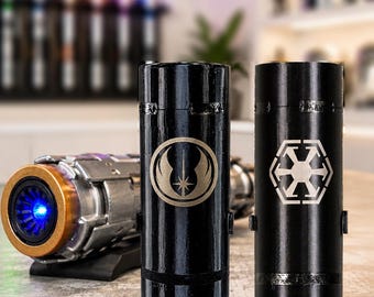 May include: Two black cylindrical containers featuring silver emblems, one with a Jedi Order symbol and the other with a Galactic Empire logo, positioned in front of a metallic lightsaber hilt with a glowing blue light.
