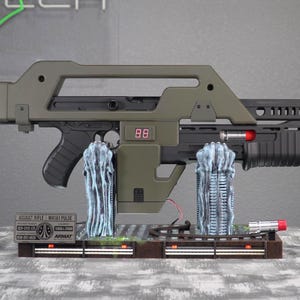 May include: A detailed replica of a sci-fi assault rifle, primarily in olive green and black, with a digital display showing "88". The model includes intricate details and is mounted on a display base with alien-like structures and a small sign.