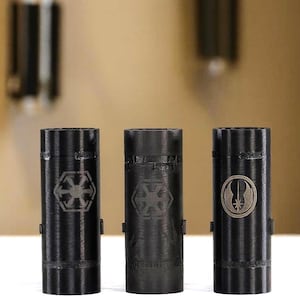 May include: Three cylindrical black objects with textured surfaces, each featuring a distinct engraved emblem. The left and center objects display a six-spoked gear-like symbol, while the right object features a circular emblem with a central star.