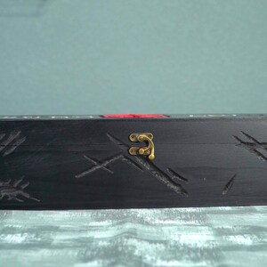 May include: A black wooden box with carved scratch marks and a brass clasp. The box has a red accent and is rectangular in shape. The box is sitting on a textured surface.