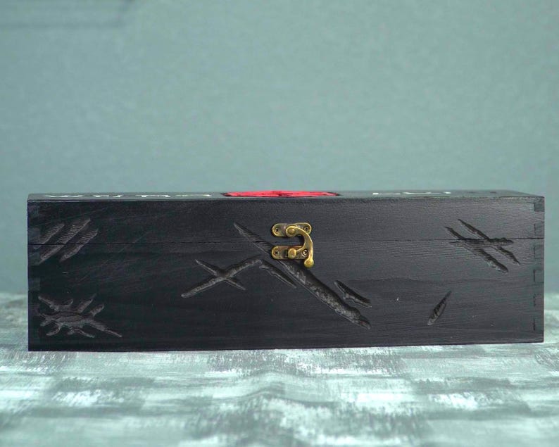 May include: A black wooden box with carved scratch marks and a brass clasp. The box has a red accent and is rectangular in shape. The box is sitting on a textured surface.
