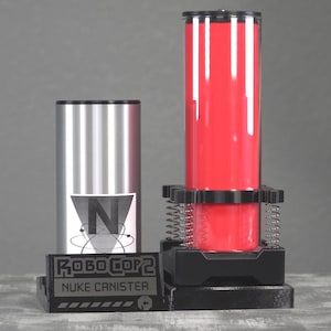 May include: A Robocop 2 Nuke Canister prop. The prop features a silver canister with a black top, a red canister with a black top and base, and a black base with the text "ROBOCOP 2 NUKE CANISTER".