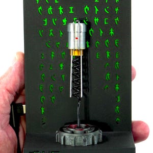 May include: A black display featuring green text and symbols from The Matrix. A silver and gold object with a red accent is suspended in front of the display. The base of the display reads "THE MATRIX" in green.