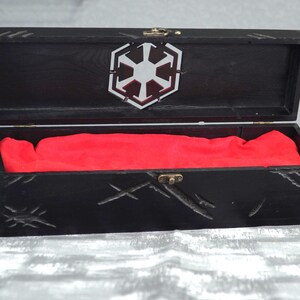 May include: Black wooden box with a hinged lid, featuring a silver emblem and red fabric lining. The box has carved details and a brass clasp. The open box reveals the red interior.