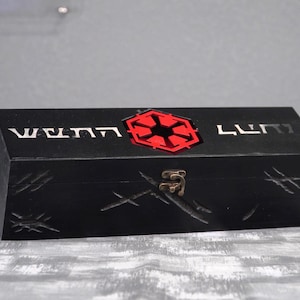 May include: A black wooden box with a red and black emblem on the top. The box has etched designs and the text "WENN LUMI" in silver. A metal clasp secures the box.