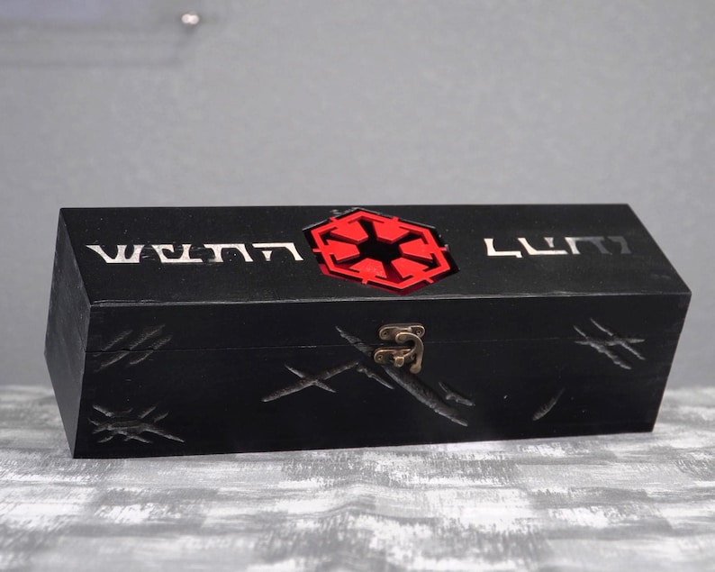 May include: A black wooden box with a red and black emblem on the top. The box has etched designs and the text "WENN LUMI" in silver. A metal clasp secures the box.