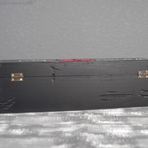 May include: A black wooden box with brass hinges and a red accent. The box has a distressed finish, with visible wood grain and imperfections. The box is rectangular and appears to be designed for storage or display.