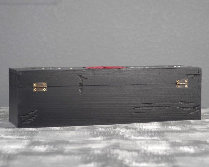 May include: A black wooden box with brass hinges and a red accent. The box has a distressed finish, with visible wood grain and imperfections. The box is rectangular and appears to be designed for storage or display.