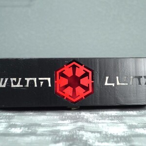 May include: A black rectangular box with silver lettering and a red illuminated emblem. The emblem is a six-sided shape with a starburst design in the center. The box has a textured surface.