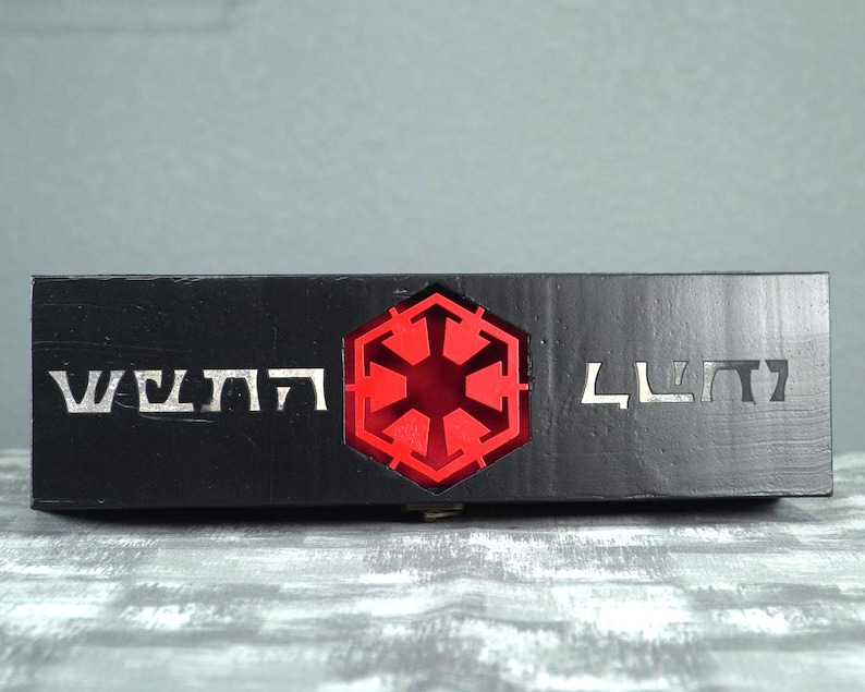 May include: A black rectangular box with silver lettering and a red illuminated emblem. The emblem is a six-sided shape with a starburst design in the center. The box has a textured surface.