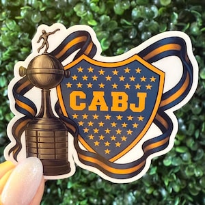 May include: A sticker featuring the CABJ logo, a shield with gold stars and the letters CABJ in gold. The sticker also includes a trophy and a blue and gold ribbon design.