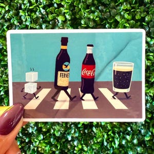 May include: A sticker featuring an illustration of a crosswalk with a bottle of Fernet, a Coca-Cola bottle, a glass of dark liquid, and two sugar cubes walking across it. The sticker is held by a hand with a maroon nail polish.