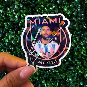 May include: A holographic sticker featuring a portrait of a soccer player. The sticker has the words "MIAMI" and "MESSI" in white text. The background is a green, leafy texture.