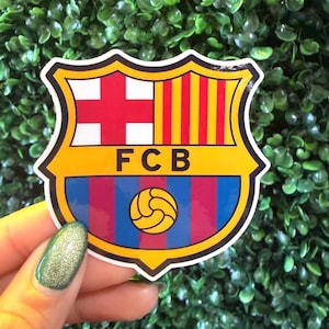 May include: A sticker featuring the FC Barcelona crest. The crest is shield-shaped with a red cross on a white background, vertical yellow and red stripes, and the letters "FCB". A soccer ball is at the bottom.