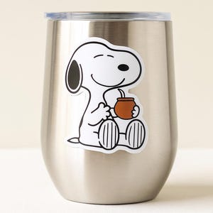 May include: A stainless steel insulated tumbler with a Snoopy sticker. The sticker shows Snoopy sitting and holding a brown cup with a straw. The tumbler has a silver lid and a curved body.