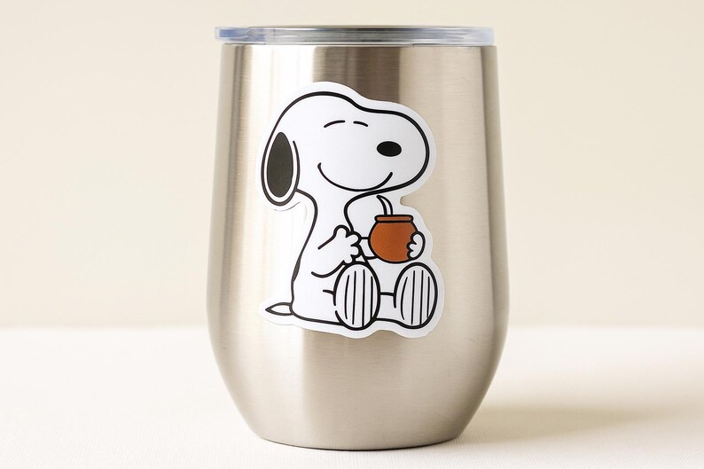 May include: A stainless steel insulated tumbler with a Snoopy sticker. The sticker shows Snoopy sitting and holding a brown cup with a straw. The tumbler has a silver lid and a curved body.