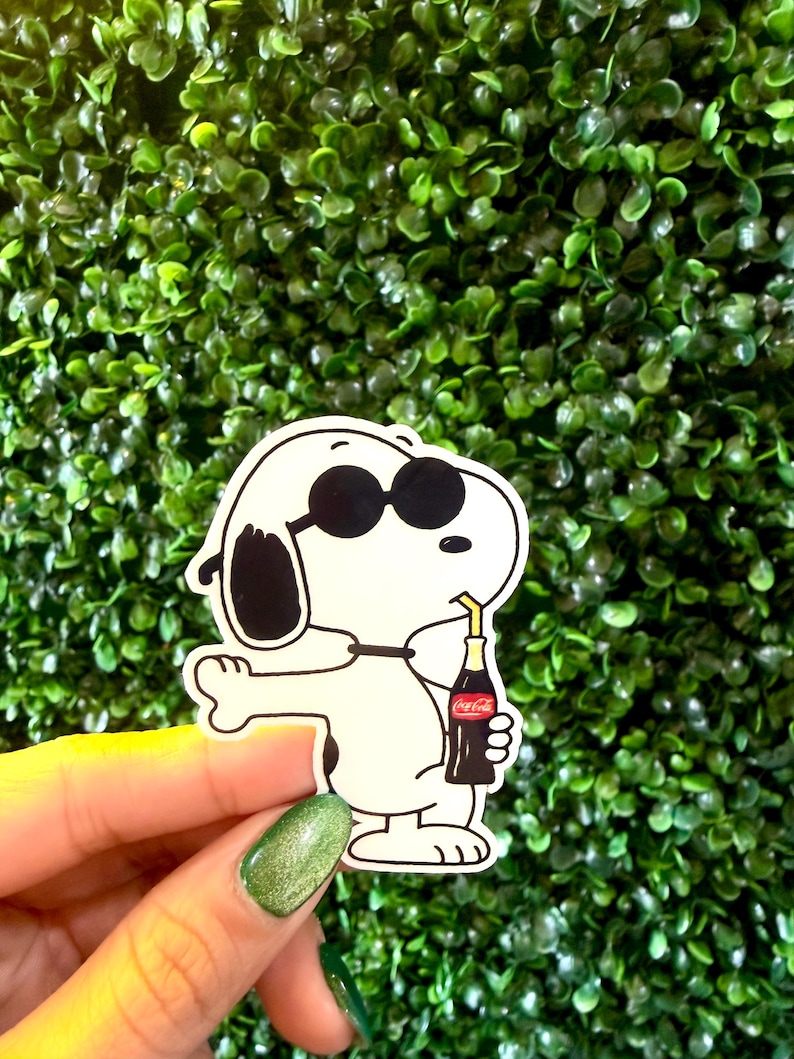 K&ouml;nnte beinhalten: Ein wei&szlig;er Snoopy-Aufkleber mit Sonnenbrille, der eine Coca-Cola-Flasche mit Strohhalm h&auml;lt. Der Aufkleber hat schwarze Akzente und befindet sich vor einem gr&uuml;nen Hintergrund.