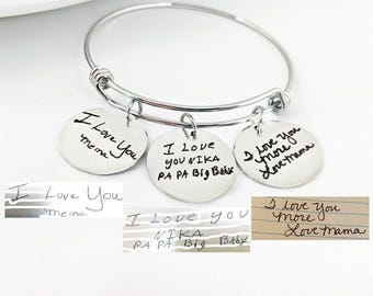 Puede incluir: Una pulsera de brazalete plateada con tres colgantes redondos. Cada colgante tiene texto escrito a mano: "I Love You Mema", "I Love You Nika Pa Pa Big Baby", y "I Love You More Love Mama".