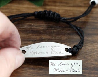May include: A silver bracelet with a black adjustable cord. The bracelet has an oval metal plate engraved with the words "We love you, Mom + Dad." A handwritten note with the same text is also visible.