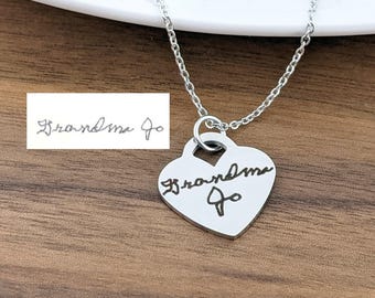 Puede incluir: Un collar con colgante en forma de corazón plateado con el texto manuscrito "Grandma Jo". El collar está sobre una cadena plateada. Una pequeña imagen rectangular muestra el mismo texto.