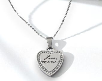 Puede incluir: Un collar con colgante en forma de corazón plateado. El colgante tiene un borde con cuentas y el texto manuscrito "Love, Mama" en negro. La cadena del collar también es plateada.