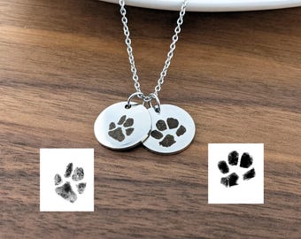 Op de afbeelding: Een zilveren ketting met twee ronde hangers met poot afdrukken. De poot afdrukken zijn zwart en lijken van een hond te zijn.