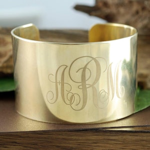 Puede incluir: Un brazalete ancho de color dorado con iniciales grabadas. El brazalete tiene una superficie lisa y pulida y un diseño sencillo y abierto. Las iniciales están en una fuente cursiva decorativa. Una joya elegante.