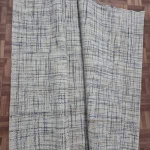 May include: A rectangular woven rug with a neutral color palette. The rug features a textured, grid-like pattern in shades of beige, gray, and brown. The rug is partially rolled up, revealing its thickness and construction. The rug is suitable for home decor.