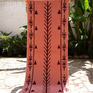 May include: A rectangular, coral-colored rug with a geometric pattern. The design features a central vertical line with branching elements and triangles, flanked by lines with stylized leaf shapes. The rug is held up outdoors.