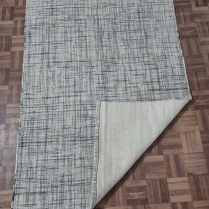 May include: A rectangular area rug with a woven, textured design in shades of gray and blue. The rug's underside is a solid, light beige color, with a corner folded up to reveal the backing. The rug is placed on a wooden floor.