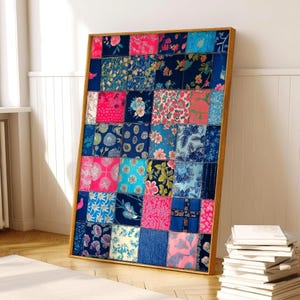 May include: A framed patchwork textile art piece. The artwork features a grid of fabric squares in various shades of blue, pink, and floral patterns. The frame is made of light-colored wood, and the piece is leaning against a white wall.
