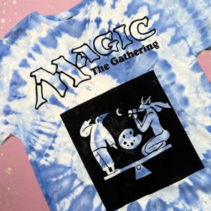 May include: Tie-dye t-shirt with a blue and white pattern. The shirt features the text "MAGIC The Gathering" in black. A black square graphic on the shirt depicts two figures on a seesaw, one with a palette.