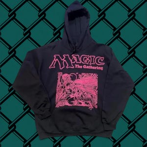 May include: A black hooded sweatshirt featuring the pink text Magic The Gathering and a pink graphic illustration of a winged figure in a forest setting on the front. The garment includes a hood with drawstrings and a large front kangaroo pocket.