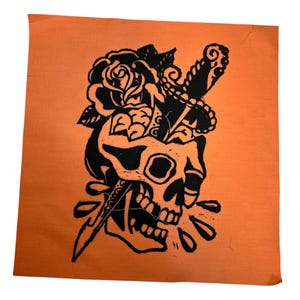 Puede incluir: Textil cuadrado naranja con un diseño de calavera negra. La calavera tiene una rosa y una daga atravesándola. El diseño es un grabado en linóleo.