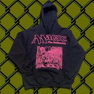 May include: A black hooded sweatshirt featuring the pink text Magic The Gathering and a pink graphic illustration of a winged figure kneeling in a forest setting. The garment includes a front kangaroo pocket, long sleeves, and a drawstring hood.