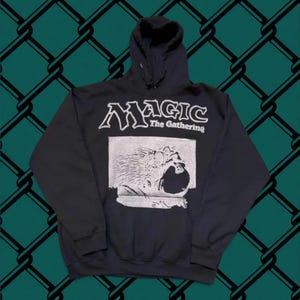 May include: A black long-sleeve hooded sweatshirt featuring the white text Magic The Gathering at the top. Below the text is a rectangular graphic print in white depicting a stylized creature with textured details on a solid black background.