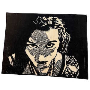 Puede incluir: Impresión artística gráfica en blanco y negro de un rostro con mirada intensa y un collar. El estilo artístico presenta sombreado y líneas audaces, creando un contraste llamativo. La impresión está sobre tela rectangular.