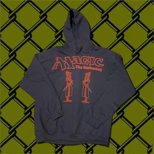 May include: A dark gray hooded sweatshirt featuring the orange text Magic The Gathering and a graphic illustration of two figures standing face to face. The hoodie includes long sleeves, a front kangaroo pocket, and a drawstring hood.