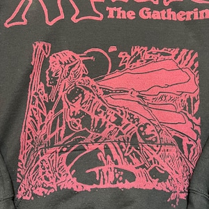 Puede incluir: Una sudadera con capucha negra con un estampado gr&aacute;fico rojo. El texto "MAGIC The Gathering" est&aacute; impreso en rojo en la parte superior. Debajo del texto hay una ilustraci&oacute;n roja detallada de una figura con orejas puntiagudas en un bosque.