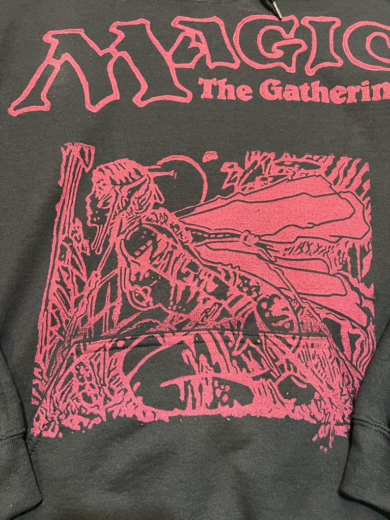 Puede incluir: Una sudadera con capucha negra con un estampado gr&aacute;fico rojo. El texto "MAGIC The Gathering" est&aacute; impreso en rojo en la parte superior. Debajo del texto hay una ilustraci&oacute;n roja detallada de una figura con orejas puntiagudas en un bosque.