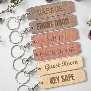 Könnte beinhalten: Holz-Schlüsselanhänger mit silberfarbenen Verschlüssen. Jeder Schlüsselanhänger ist mit einem anderen Ort graviert: "GARAGE", "FRONT DOOR", "SHED", "BACK DOOR", "Guest Room" und "KEY SAFE". Die Schlüsselanhänger sind in verschiedenen Brauntönen gehalten.